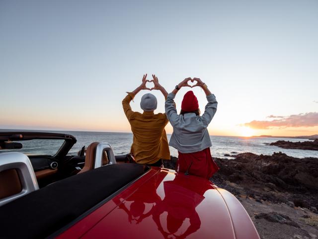 Couple enjoying beautiful views on the ocean, standing together near the car on the rocky coast, showing with hands heart shape. Carefree lifestyle, love and travel concept